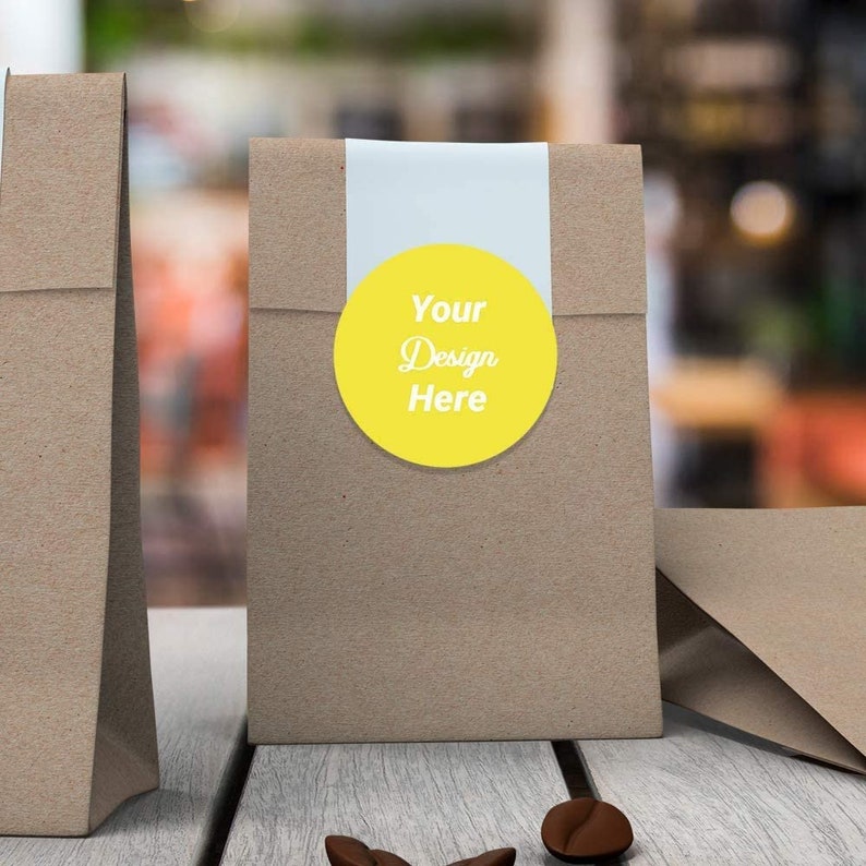 Pu&ograve; includere: Due sacchetti di carta marrone con un adesivo circolare giallo su uno dei sacchetti. L'adesivo ha il testo "Your Design Here".