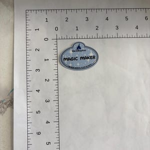 May include: An oval-shaped Walt Disney World name tag sticker with a light blue background and white star accents. The text reads MAGIC MAKER and THE MOST MAGICAL PLACE ON EARTH. It is positioned next to an L-shaped ruler showing a width of 5.08 cm.