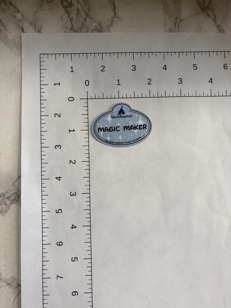 May include: An oval-shaped Walt Disney World name tag sticker with a light blue background and white star accents. The text reads MAGIC MAKER and THE MOST MAGICAL PLACE ON EARTH. It is positioned next to an L-shaped ruler showing a width of 5.08 cm.
