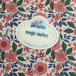 May include: An oval-shaped Disneyland name tag featuring the text magic maker and The Happiest Place on Earth, set against a white background with a repeating pattern of pink flowers, dark green leaves, and small orange berries.