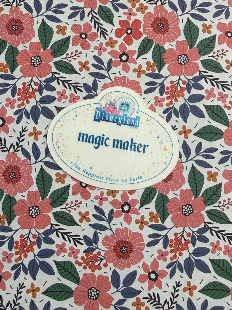 May include: An oval-shaped Disneyland name tag featuring the text magic maker and The Happiest Place on Earth, set against a white background with a repeating pattern of pink flowers, dark green leaves, and small orange berries.
