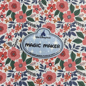 May include: An oval-shaped Walt Disney World name tag featuring the text MAGIC MAKER and THE MOST MAGICAL PLACE ON EARTH, set against a white background with a repeating pattern of pink flowers, dark green leaves, and small orange berries.
