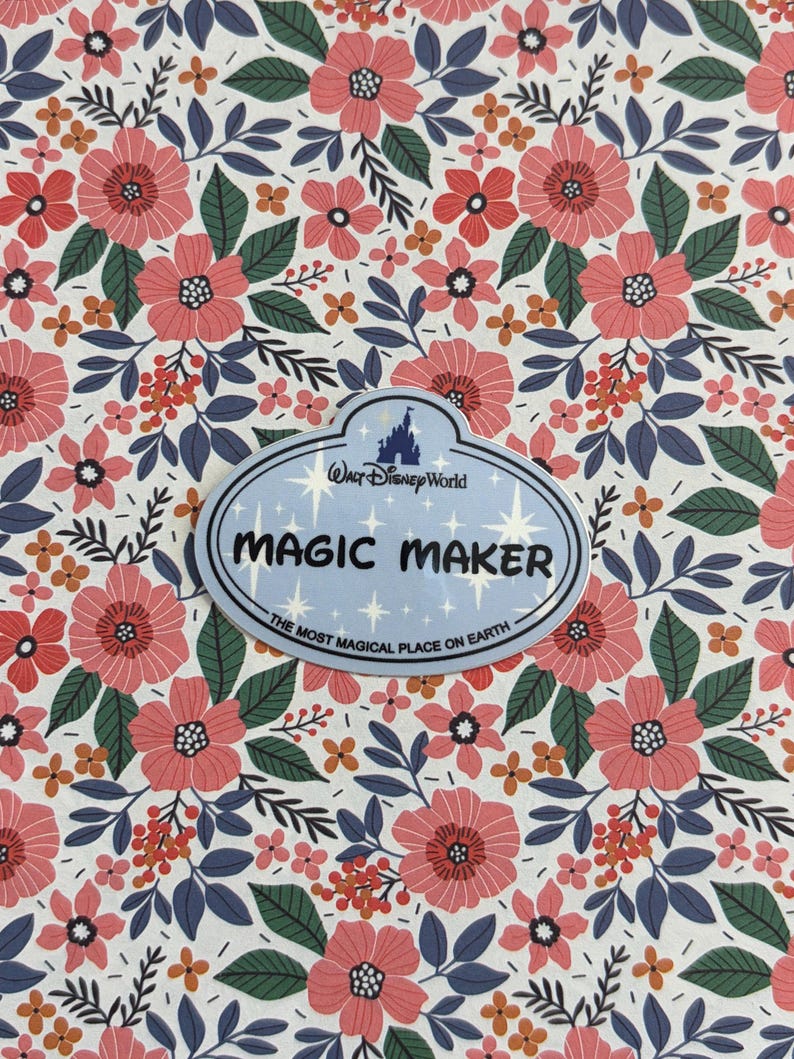May include: An oval-shaped Walt Disney World name tag featuring the text MAGIC MAKER and THE MOST MAGICAL PLACE ON EARTH, set against a white background with a repeating pattern of pink flowers, dark green leaves, and small orange berries.