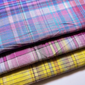 May include: Three folded layers of woven fabric featuring a plaid pattern. The top layer displays blue, purple, and pink tones. The middle layer shows magenta, white, and grey. The bottom layer features a bright yellow base with brown and white lines.
