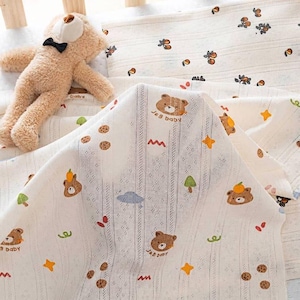 May include: A light beige teddy bear with a black bow tie sits next to a white cotton fabric featuring a repeating pattern of brown bear faces, orange fruit, green trees, and yellow stars, with the text J&B baby printed near the bear faces.