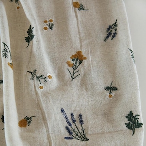 May include: A light beige fabric featuring scattered embroidery of various wildflowers and herbs. The design includes small yellow blossoms, white daisy-like flowers, dark green leafy sprigs, and clusters of purple lavender-colored stems.