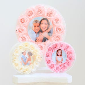 May include: Three circular photo frames with floral rose accents. The largest frame features a photo of two people, while the smaller frames display individual photos. The roses are in shades of pink and white.
