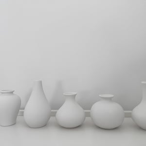 Pode incluir: Cinco vasos de cerâmica branca de diferentes formas e tamanhos dispostos em uma fileira sobre uma superfície branca.