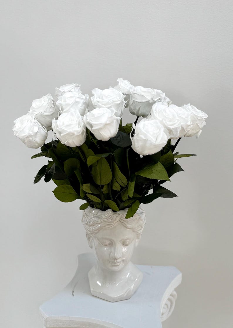 May include: A white ceramic bust vase with a bouquet of white roses. The vase is shaped like a woman's head with a crown of leaves.