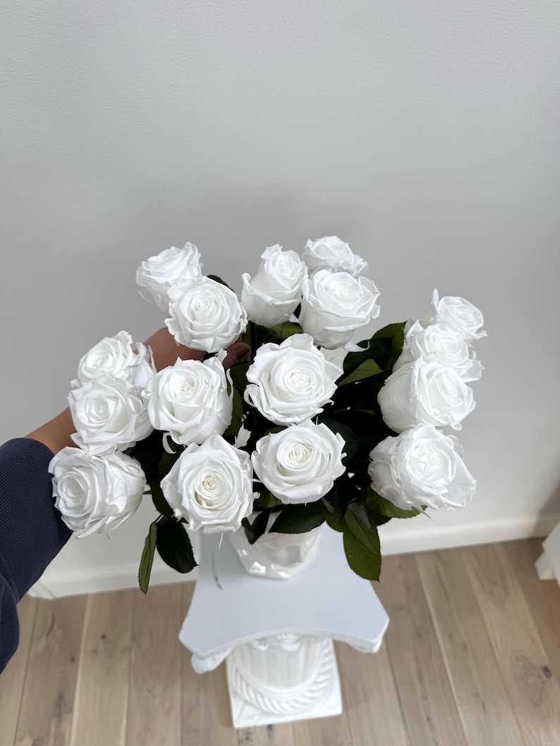 May include: A bouquet of white roses in a clear glass vase, arranged on a white pedestal.