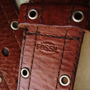 May include: Close-up of a brown leather belt with metal eyelets and stitching. The embossed text reads "GENUINE FOSSIL ORIGINAL". The belt is a rich, warm brown color, suggesting a vintage or rustic style.