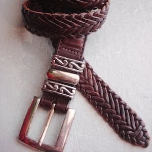 May include: A brown leather braided belt with a silver buckle. The buckle has a decorative design.