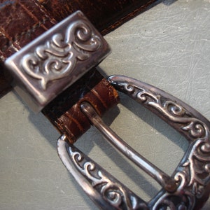 May include: A brown leather belt with a silver buckle featuring an ornate design. The buckle is rectangular with a decorative border and a floral pattern.
