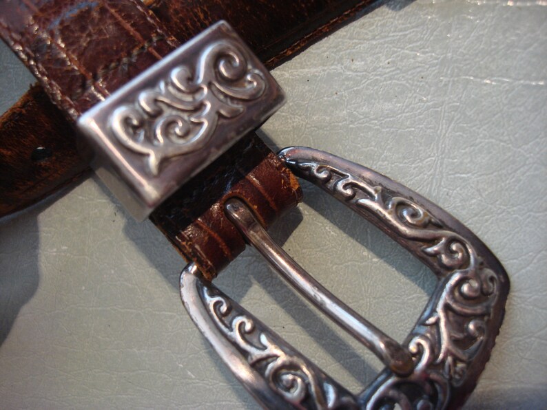 May include: A brown leather belt with a silver buckle featuring an ornate design. The buckle is rectangular with a decorative border and a floral pattern.