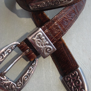 May include: A brown leather belt with a silver buckle and decorative accents. The buckle has a floral design and the belt has a textured surface.