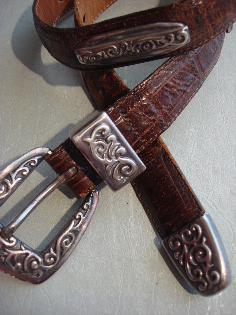 May include: A brown leather belt with a silver buckle and decorative accents. The buckle has a floral design and the belt has a textured surface.