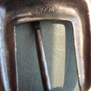 May include: Close-up of a silver metal belt buckle with the inscription "&copy; 1994".