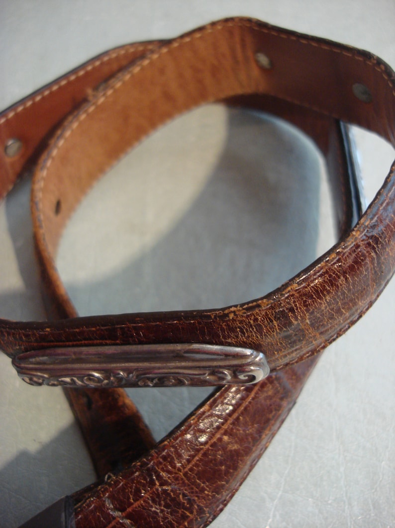 May include: A brown leather belt with a silver buckle. The buckle has a decorative design.