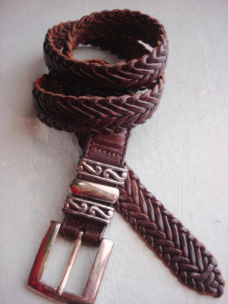 May include: A brown leather braided belt with a silver buckle. The buckle has a decorative design.