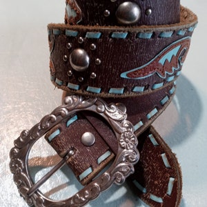 May include: A brown leather belt with a silver buckle and turquoise stitching. The belt has decorative metal accents and a southwestern design.