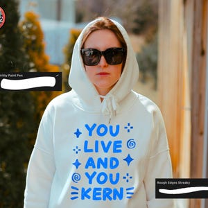 May include: A person wearing a white hooded sweatshirt with the text "YOU LIVE AND YOU KERN" in blue. The person is also wearing black sunglasses. The image has a warm, natural light.