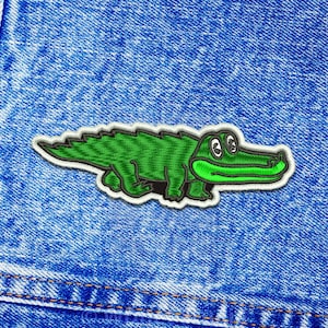 Peut inclure: Patch brodé d'un alligator vert sur un fond en jean bleu.