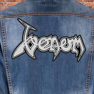 May include: A denim jacket with a large, white and black Venom patch on the back. The patch features the band's name in a stylized, gothic font. The jacket is a light blue wash.