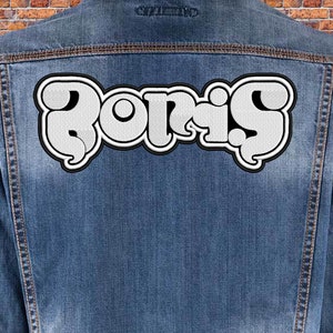 Puede incluir: Chaqueta vaquera azul con el nombre "BORIS" bordado en la espalda. Las letras blancas están delineadas en negro. El fondo es una pared de ladrillos.