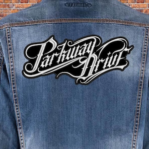 Könnte beinhalten: Ein gestickter schwarzer und weißer Aufnäher mit dem Text "Parkway Drive" in einer stilisierten Schriftart. Der Aufnäher ist auf eine blaue Jeansjacke genäht.