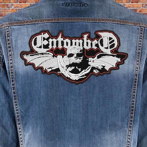 May include: A black and white embroidered patch featuring the band name "Entombed" in a gothic font. The patch depicts a skull with wings and a chain around its neck.