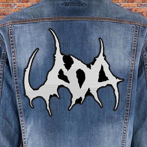 以下が含まれることがあります： 背面に白と黒の刺繍パッチが付いたデニムジャケット。パッチは、ゴシック体で「GOD」とスタイリッシュに書かれたロゴです。