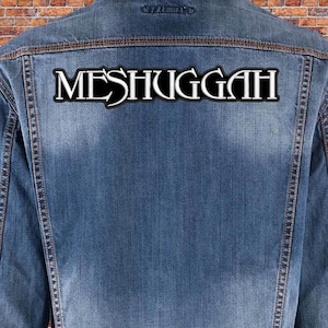 Op de afbeelding: Een blauw spijkerjack met een grote, rechthoekige geborduurde patch op de achterkant. De patch heeft een zwarte achtergrond met een witte rand en het woord MESHUGGAH staat in een gestileerd, vet wit lettertype in het midden van de stof.