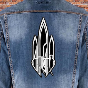 Op de afbeelding: Een zwart-witte patch met het woord "At The Gates" in een gotische lettertype. De patch is op een blauwe denim jas.