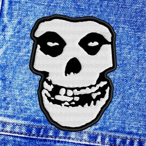 Può includere: Toppa ricamata bianca a forma di teschio con occhi neri e un ghigno beffardo, su uno sfondo di denim blu.