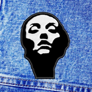 Peut inclure: Écusson brodé noir et blanc représentant un visage stylisé. Le visage est blanc avec des détails noirs, sur un fond noir. L'écusson est cousu sur un tissu denim bleu, visible en arrière-plan.