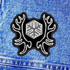 May include: Black and white embroidered patch with a snowflake design inside a hexagonal shape, surrounded by two stylized antlers. The patch is sewn onto a blue denim fabric.