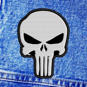 May include: Embroidered patch depicting a white skull with black eye sockets and a black border. The skull has a vertical, toothed jawline. The patch is set against a blue denim background, with visible stitching.