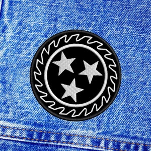 May include: Black and white embroidered patch with three white stars inside a circle with a sawtooth border. The patch is on a blue denim background.