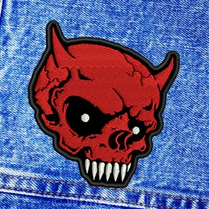 May include: Red devil skull embroidered patch with black eyes and white teeth. The patch is on a blue denim background.