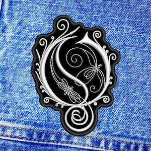 May include: A black and white embroidered patch featuring a stylized letter G with intricate swirling filigree and decorative line work. The patch has a black border and is centered on a textured blue denim fabric background with visible stitching.