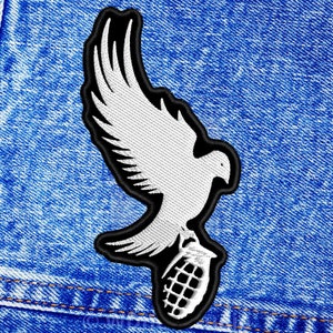 Könnte beinhalten: Eine weiße Taube mit schwarzem Umriss, die eine Granate in ihren Krallen hält. Die Taube fliegt mit ausgebreiteten Flügeln. Das Bild ist auf einem blauen Jeans-Hintergrund.