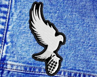 May include: A white dove with a black outline holding a grenade in its talons. The dove is flying with its wings spread out. The image is on a blue denim background.