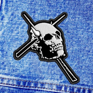 Op de afbeelding: Een geborduurde patch in zwart en wit van een schedel met twee gekruiste drumsticks. De schedel heeft een witte omtrek en de drumsticks zijn zwart.