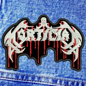 May include: An embroidered patch featuring the word Mortician in a stylised white font with a skull icon above the text. The design includes red vertical drips against a black background, all outlined in black, attached to blue denim fabric.