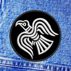 Könnte beinhalten: Ein gestickter schwarzer und weißer Aufnäher mit einem stilisierten Raben auf schwarzem Hintergrund. Der Aufnäher ist auf einem blauen Jeansstoff genäht.