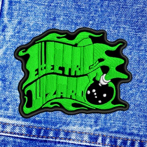 Könnte beinhalten: Gestickter Aufnäher mit den Worten "ELECTRIC WIZARD" in grüner, stilisierter Schrift. Der Aufnäher hat einen schwarzen Rand und ein grünes, flüssigkeitsartiges Design um den Text. Der Aufnäher ist vor einem blauen Jeanshintergrund platziert.