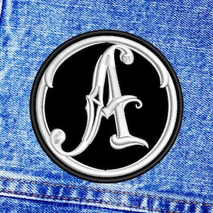 May include: A black and white embroidered patch with a stylised letter A in a circle. The patch is on a blue denim background.