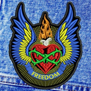 Puede incluir: Un parche bordado negro con un corazón rojo rodeado de espinas verdes y alas amarillas y azules. El corazón tiene un símbolo de infinito verde en su interior. El parche tiene una pancarta amarilla con la palabra "FREEDOM" en azul.