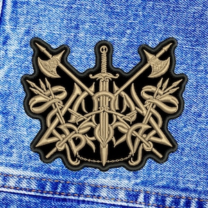 Peut inclure: Écusson brodé avec un motif symétrique de haches, d'épées et de trident croisés en fil doré sur fond noir. L'écusson est bordé de coutures noires, sur fond de denim bleu.