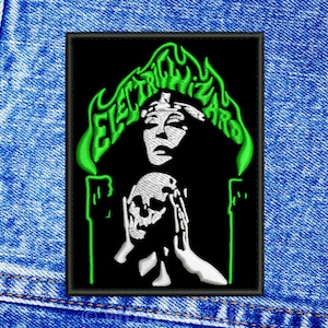Könnte beinhalten: Gestickter Aufnäher mit schwarzem Hintergrund, der ein Frauengesicht und einen Totenkopf zeigt, mit dem Text "ELECTRIC WIZARD" in grünen, flammenartigen Buchstaben. Der Aufnäher ist rechteckig und hat einen schwarzen Rand.